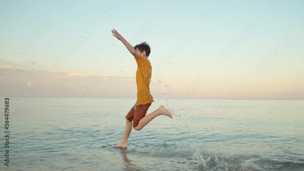 Happy boy runs in clothes jumping into sea, waving his arms, smiles splashing water around child slow motion on background of blue sky and bright sunset. Summer travel sea. Family tourism. Childhood Happy boy runs in clothes jumping into sea, waving his arms, smiles splashing water around child slow motion on background of blue sky and bright sunset. Summer travel sea. Family tourism. Childhood