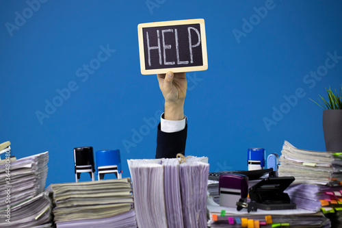 Office clerk in suit holds a blackboard with ""Help"" sign and lies under the desktop. Overload at work. Accounting paper documents piles. Isolated. Disarray in files on table. Routine paperwork