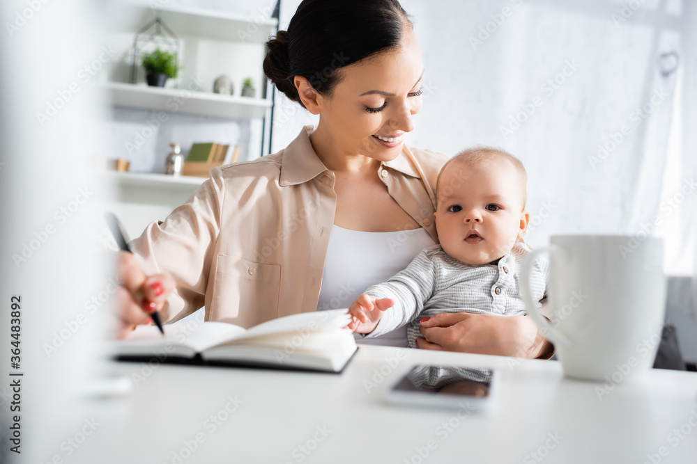 selective focus of happy mother holding pen and looking at cute infant son near smartphone selective focus of happy mother holding pen and looking at cute infant son near smartphone