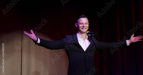 The comedian takes the stage and greets the audience against a red curtain. Portrait of young man stand up comedian speaking jokes in micropphone standing on stage. Funny performance.