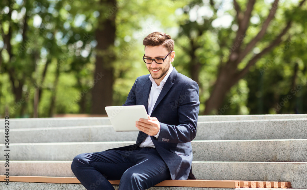 Smiling young office employee working with tablet computer while sitting on stairs at city park Smiling young office employee working with tablet computer while sitting on stairs at city park