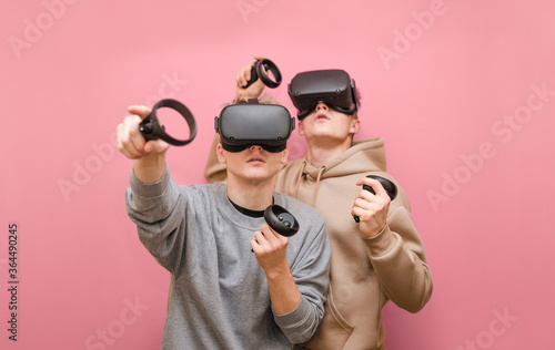 Foto Two guys in casual clothes stand on a pink background with VR helmets and play games in virtual reality