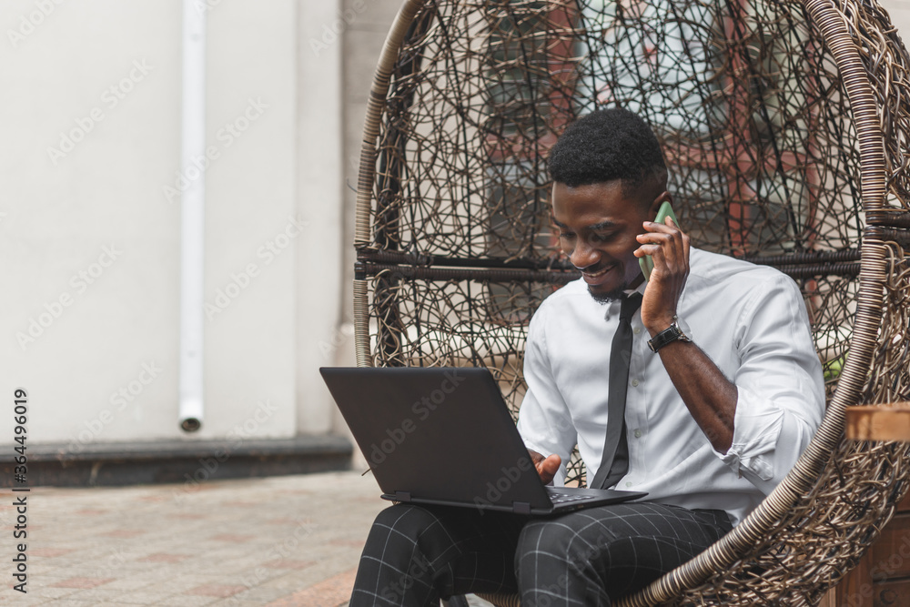 Cheerful young African man in formal wear talking on the mobile phone and working on laptop while sitting on egg chair at outdoors cafe Cheerful young African man in formal wear talking on the mobile phone and working on laptop while sitting on egg chair at outdoors cafe