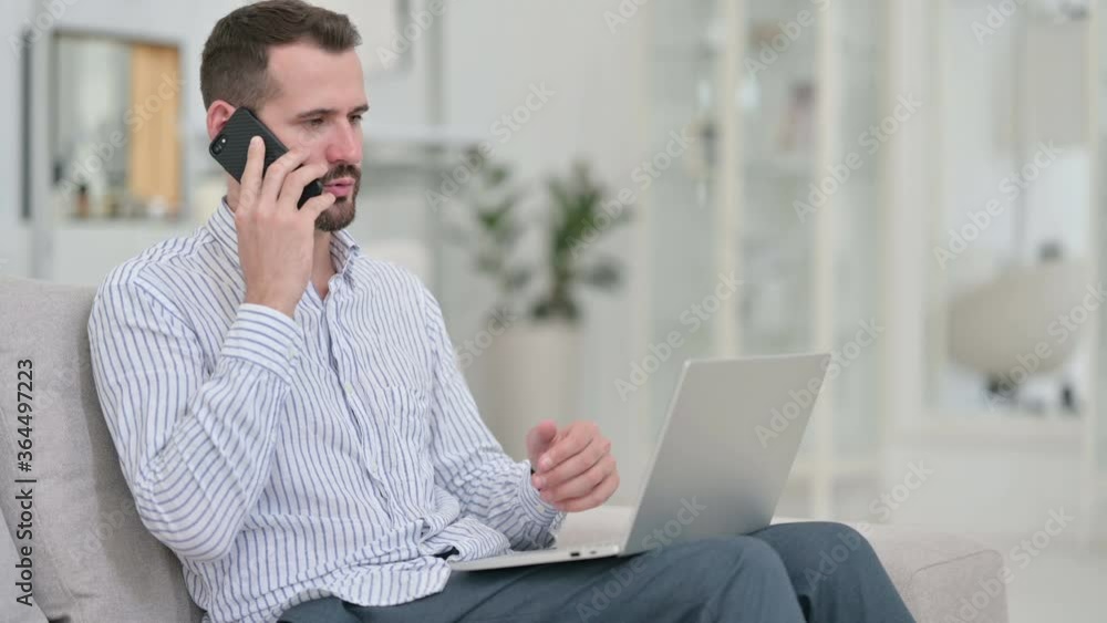 Young Man with Laptop Talking on Smartphone at Home Young Man with Laptop Talking on Smartphone at Home
