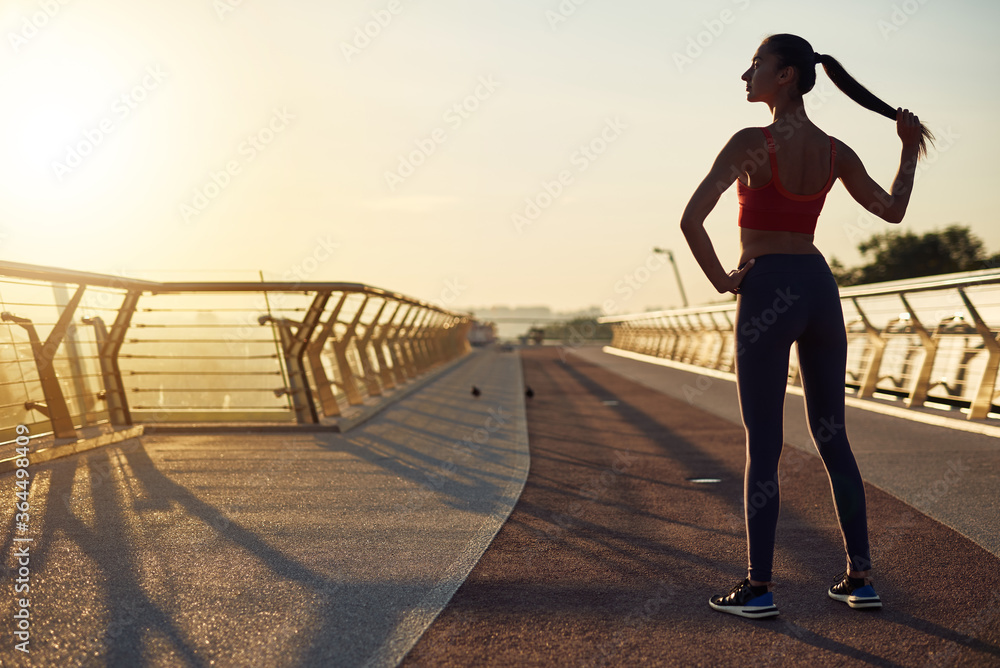 Fototapeta premium Beautiful girl working out and posing at sunrise. Young woman back view. Sport at the city. Wellness lifestyle in the city concept. Jogging in the morning