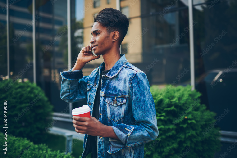 Side view of hindu young man dressed in denim jacket talking on telephone  waiting for friend on street.Stylish hipster guy with coffee to go  communicating on smartphone strolling in urban setting Stock