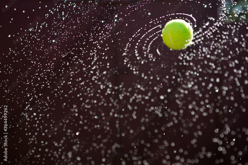 Wet Tennis ball photographed while flying and spinning at sunset on warm summer evening. Splashing water flying in different directions