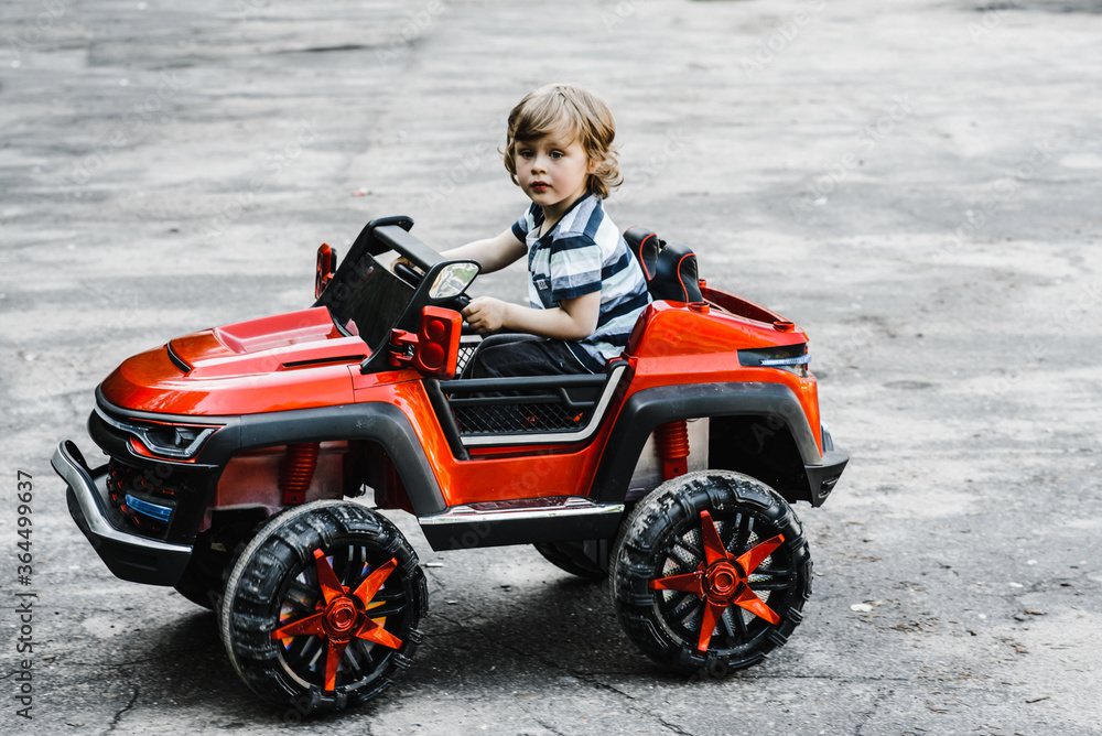 curly-haired boy in a striped T-shirt rides a red big toy car driving on an asphalt path. day off, outdoor recreation curly-haired boy in a striped T-shirt rides a red big toy car driving on an asphalt path. day off, outdoor recreation