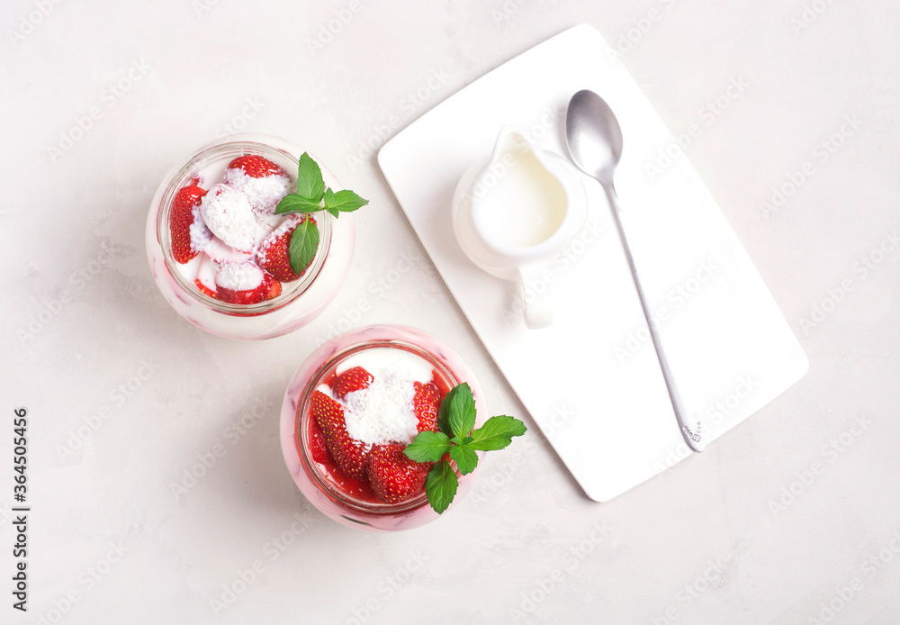 Homemade yogurt with strawberry slices, mint leaves in glass jars, next to the milkman and spoon on a gray background, top view. Healthy breakfast concept. Homemade yogurt with strawberry slices, mint leaves in glass jars, next to the milkman and spoon on a gray background, top view. Healthy breakfast concept.