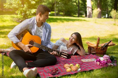 Loving man playing guitar and singing serenade to his girlfriend on romantic picnic in park