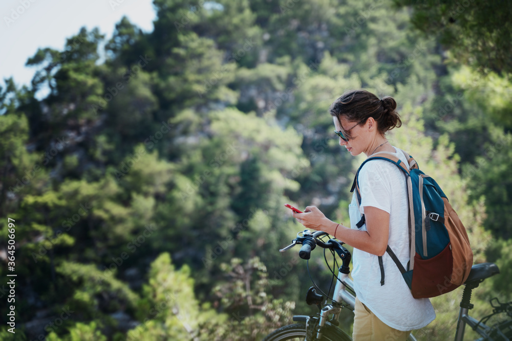 Girl with a bicycle in the forest, looks at a map on a smartphone. Summer holidays and travel by bicycle, sports and outdoor activities.
