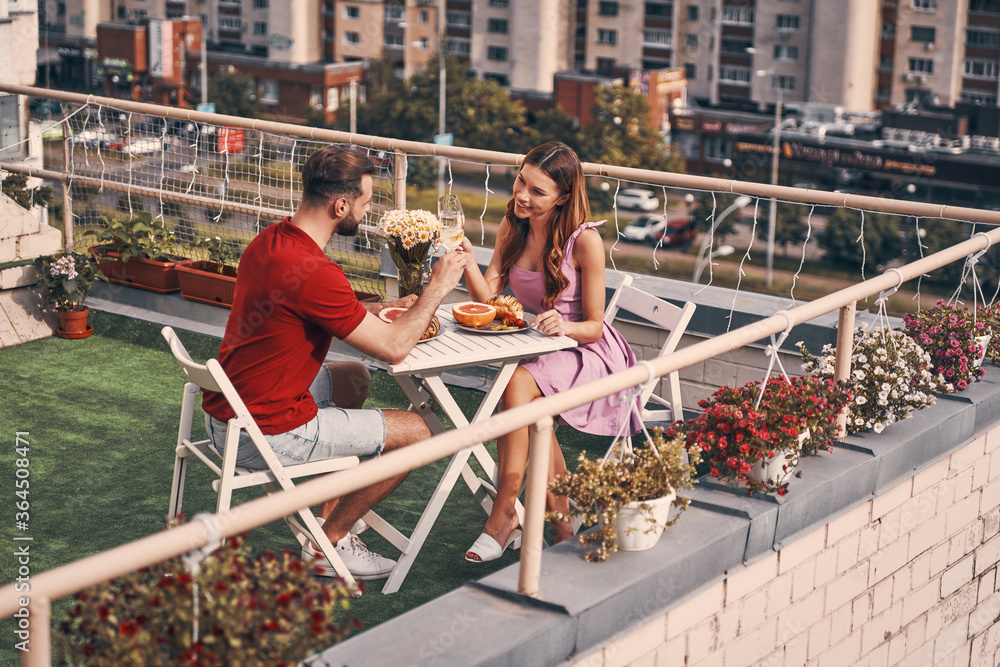 Beautiful young couple in casual clothing enjoying romantic dinner and smiling while sitting on the rooftop patio outdoors