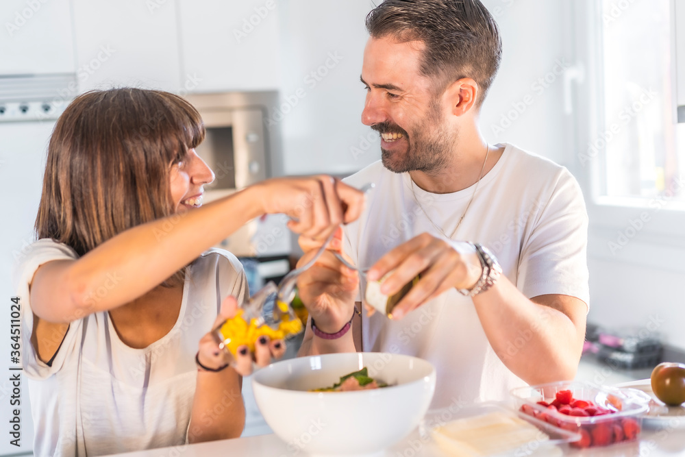 A Caucasian couple cooking together at home, cooking in confinement, cooking with the family. Coronavirus, covid-19. Putting the entrees on the salad smiling