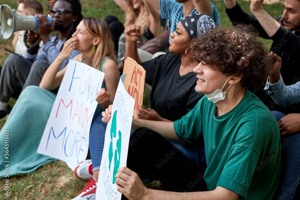 young caucasian and african american people sit on grass int he park with placards and chant slogans to demand an action on climate change young caucasian and african american people sit on grass int he park with placards and chant slogans to demand an action on climate change