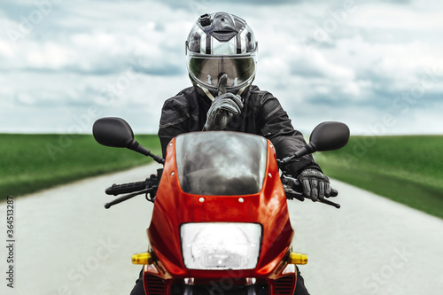 Fototapeta Naklejka Na Ścianę i Meble -  A man sits on a red motorcycle in protective gear and helmet and shows the sign quietly with his finger. Biker knows the secret.