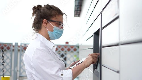 Curly woman in a mask stands near an ATM with a smartphone. Casual stylish girl in a white shirt enters a password on the parcel locker or the postamat photo