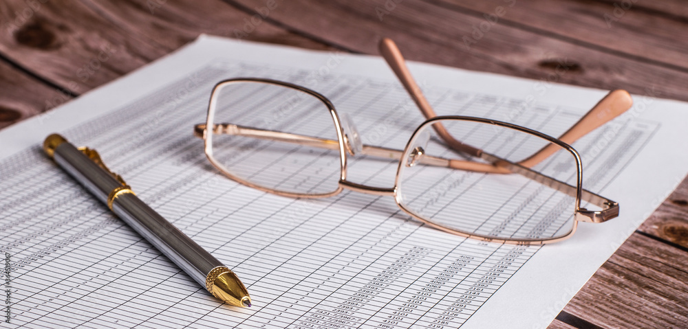 On a dark wooden table lies a document with a pen and glasses in a golden frame. Top view close up. Business concept On a dark wooden table lies a document with a pen and glasses in a golden frame. Top view close up. Business concept