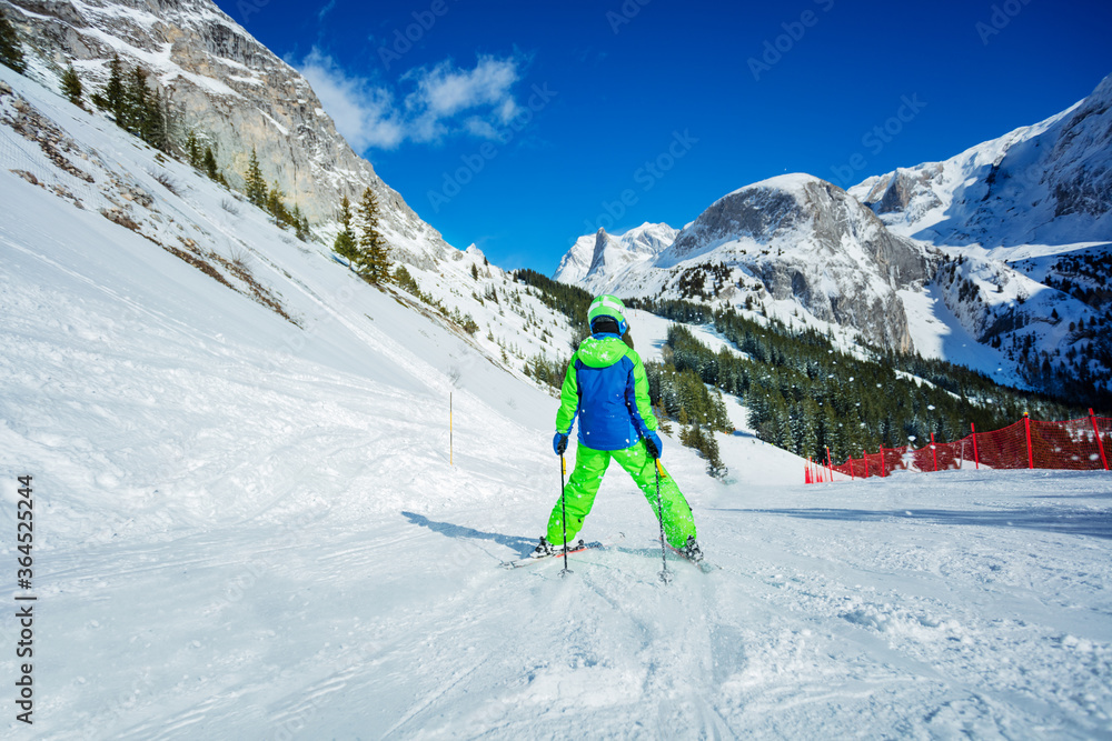 Boy skier racing down the snow track in sunny mountains wearing colorful outfit view from behind