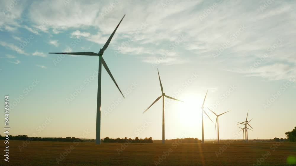 Drone shot at windmills standing in a field with wheat at sunset. Aerial view. Wind turbines rotate slowly in a beautiful field at sunset. Drone shot at windmills standing in a field with wheat at sunset. Aerial view. Wind turbines rotate slowly in a beautiful field at sunset.