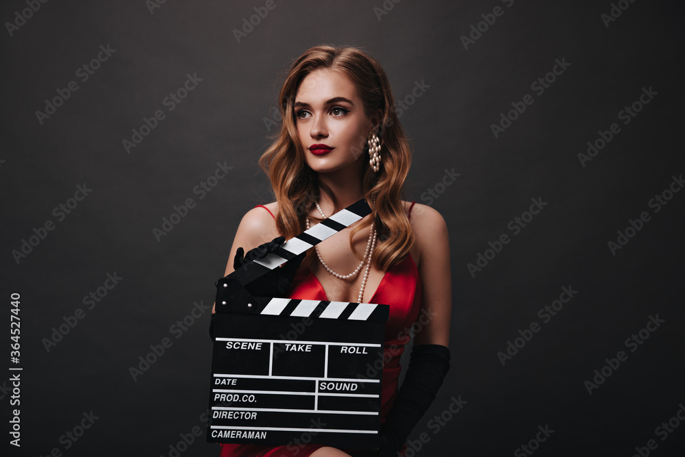 Elegant lady in beautiful jewelry holding clapperboard. Beautiful woman in red silk top posing on black isolated background