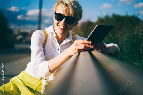 Half length portrait of positive blond young woman with stylish short haircut and trendy black sunglasses looking and smiling at camera while relaxing outdoors holding digital touch pad in hands