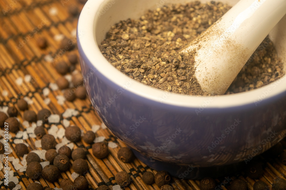 Ground black allspice in a ceramic mortar and black allspice and coarse salt scattered on a reed Mat. Close up. Ground black allspice in a ceramic mortar and black allspice and coarse salt scattered on a reed Mat. Close up.