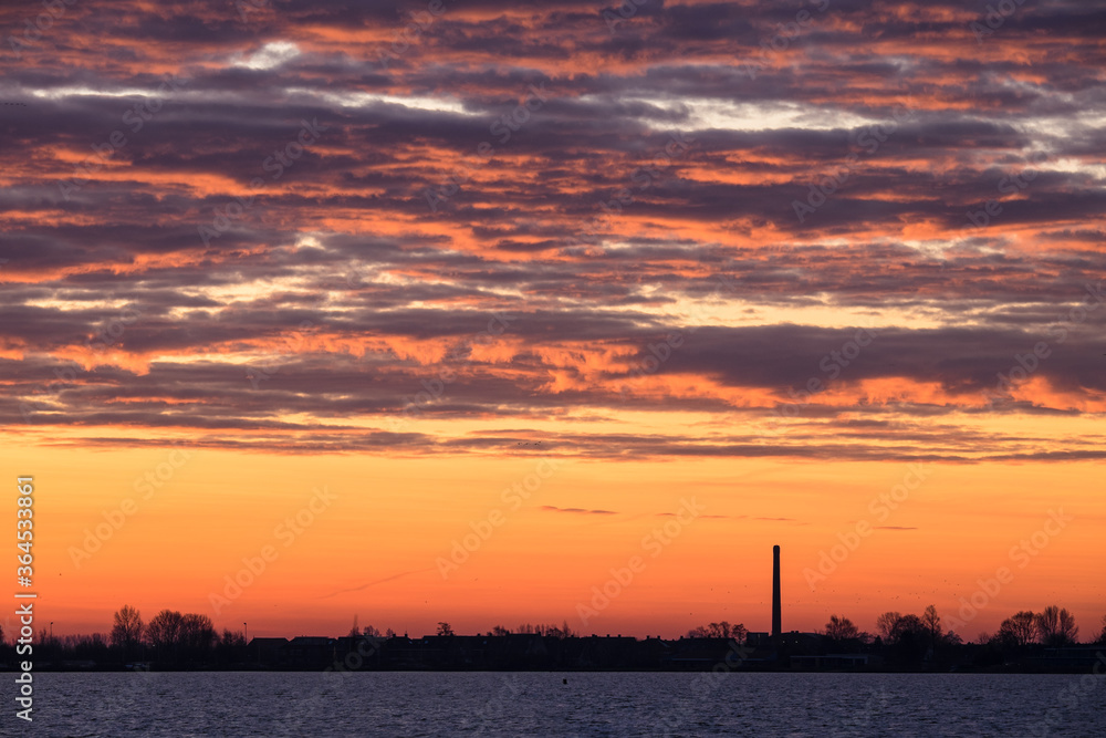 View in winter with beautiful rising sun and clouds above the Lake Tjeukemeer with chimney of a factory in the province of Friesland in the Netherlands