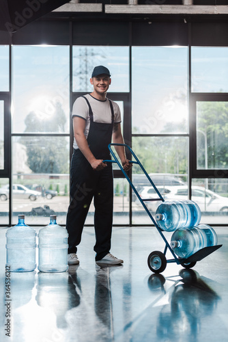 cheerful delivery man in uniform holding hand truck with bottled water