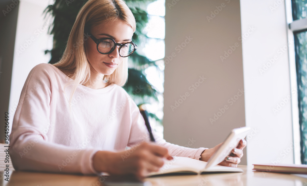 Intelligence hipster girl in eyeglasses writing information in knowledge textbook found on internet space via digital tablet, clever female student holding touchpad using for learning in school campus