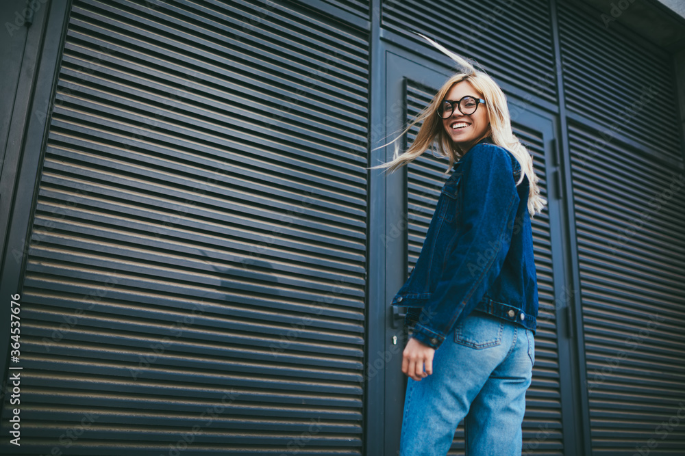 Half length portrait of happy blonde young woman in stylish eyeglasses smiling at camera.Cheerful hipster girl dressed in denim apparel while standing on promotional backgrounf with publicity area Half length portrait of happy blonde young woman in stylish eyeglasses smiling at camera.Cheerful hipster girl dressed in denim apparel while standing on promotional backgrounf with publicity area