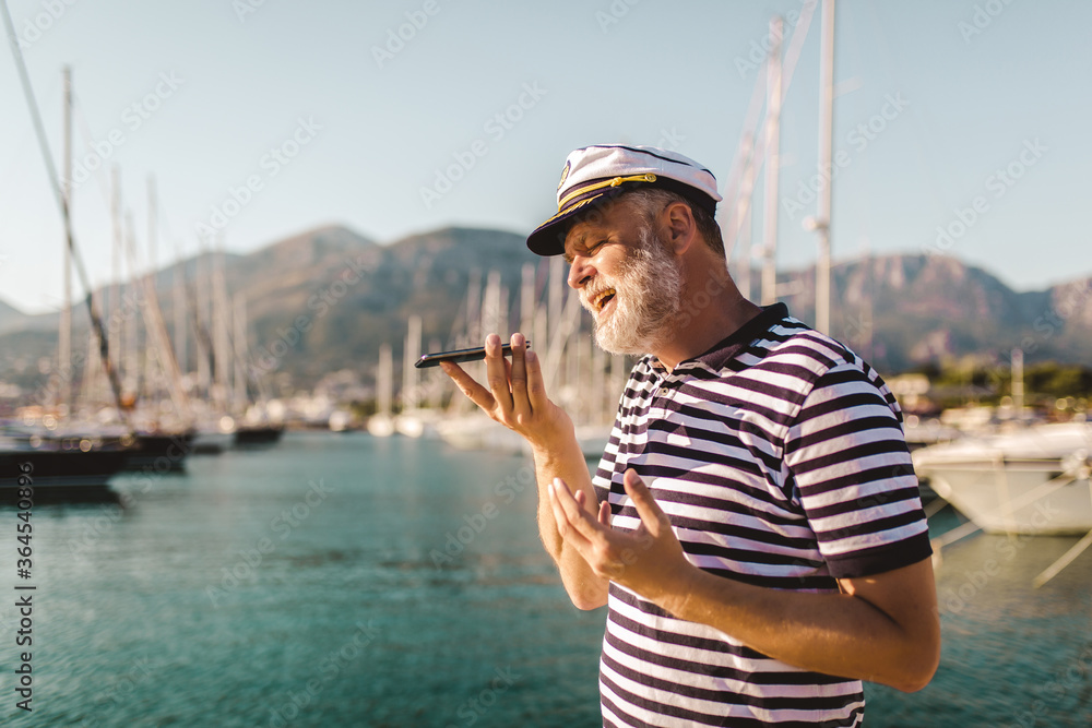 Mature man stands on pier dressed in a sailor's shirt and hat using voice command recorder on smartphone.