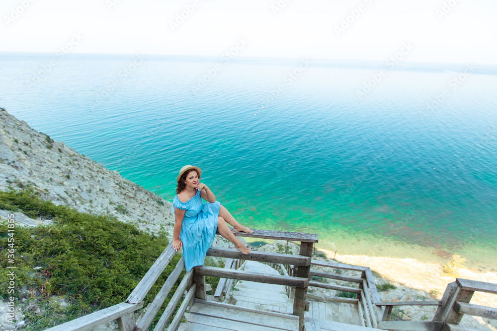 A beautiful young woman in a blue dress sits on a rock on the shore of the azure coast of the sea.