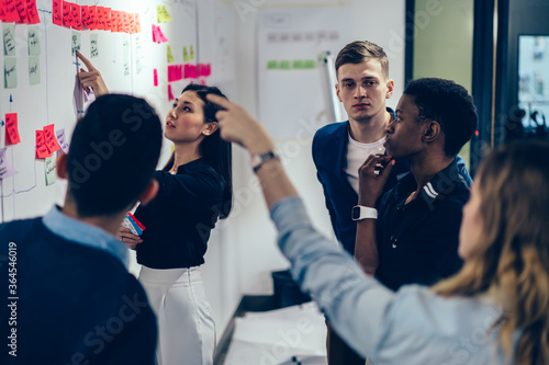 Group of professional male and female multicultural employees in formal wear pointing on glued colorful stickers on wall and teamworking in modern office on creating successful presentation