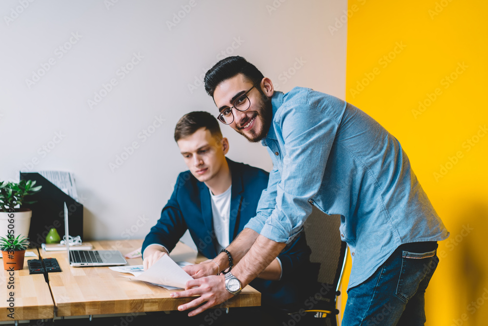 Portrait of cheerful young man smiling at camera while teamworking with colleague on developing common project.Positive male employee in formal wear consulting with proud ceo about financial reports Portrait of cheerful young man smiling at camera while teamworking with colleague on developing common project.Positive male employee in formal wear consulting with proud ceo about financial reports