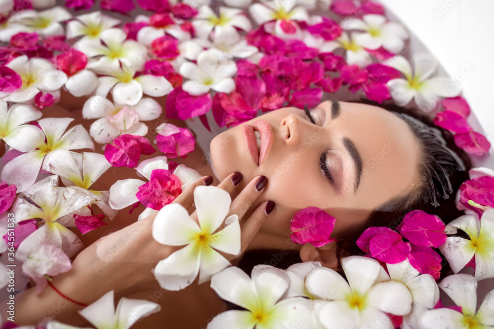 Side view. Portrait of a charming brunette with closed eyes relaxes in the bath among the flower petals. Spa and relaxation. Natural makeup