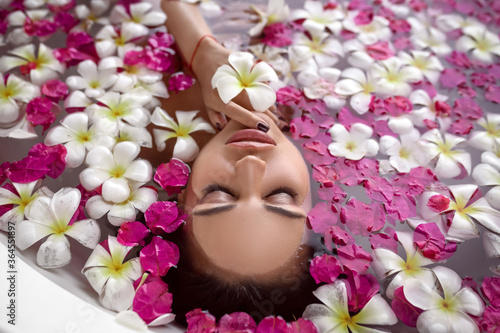 Fototapeta Naklejka Na Ścianę i Meble -  An adorable female with closed eyes relaxes in the bath among the flower pink and white petals. Spa and relaxation. Natural makeup