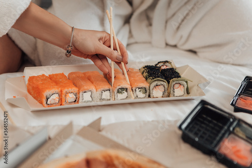 Close up shot of girl takes sushi with chopsticks. Japanese food set on eco frienly cardboard plate. Lunch break after distance work. Delivery restaurant food concept. Ladie eating ordered rolls. photo