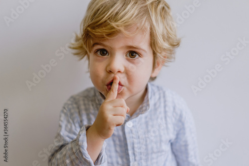 Cute little kid in casual clothing standing on white background of studio and putting index finger on lips while looking at camera photo