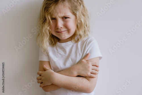 Naughty kid with wavy hair in casual clothing standing with folded arms and weeping looking at camera on white background photo