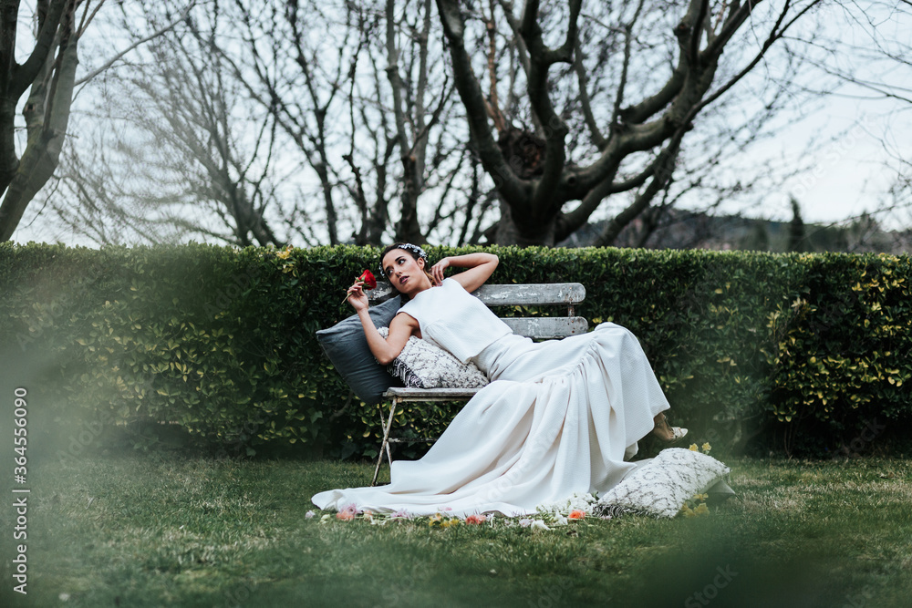 Attractive young lady in white dress and with red rose leaning on soft pillows and looking away while resting on shabby bench on wedding day in park Attractive young lady in white dress and with red rose leaning on soft pillows and looking away while resting on shabby bench on wedding day in park