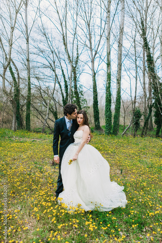 Happy groom in elegant suit hugging smiling bride with yellow dandelions on blooming meadow during wedding day Happy groom in elegant suit hugging smiling bride with yellow dandelions on blooming meadow during wedding day