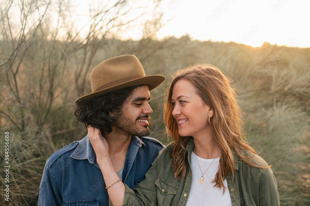 Cheerful man and woman embracing and looking at each other while standing near leafless bush during date in evening in nature Cheerful man and woman embracing and looking at each other while standing near leafless bush during date in evening in nature