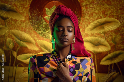 Portrait of a young african woman with closed eyes in ethnic clothes. Red scarf on the head, gold earrings. Blurred background with gold ornaments. Fashion photo