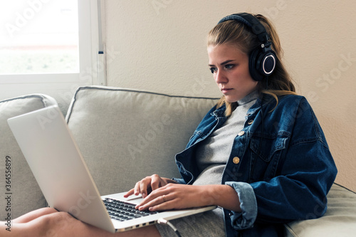 Focused young female freelancer in casual wear with wireless headphones sitting on sofa and using laptop while working on remote project at home photo