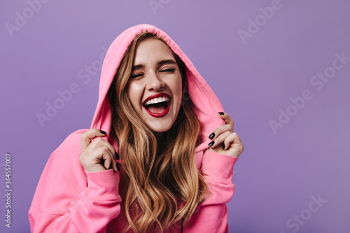 Cheerful woman in pink hoodie laughing on isolated background. Emotional happy lady in bright sweatshirt smiling on purple backdrop