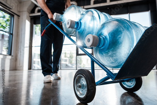 cropped view of delivery man in uniform holding hand truck with purified water in bottles