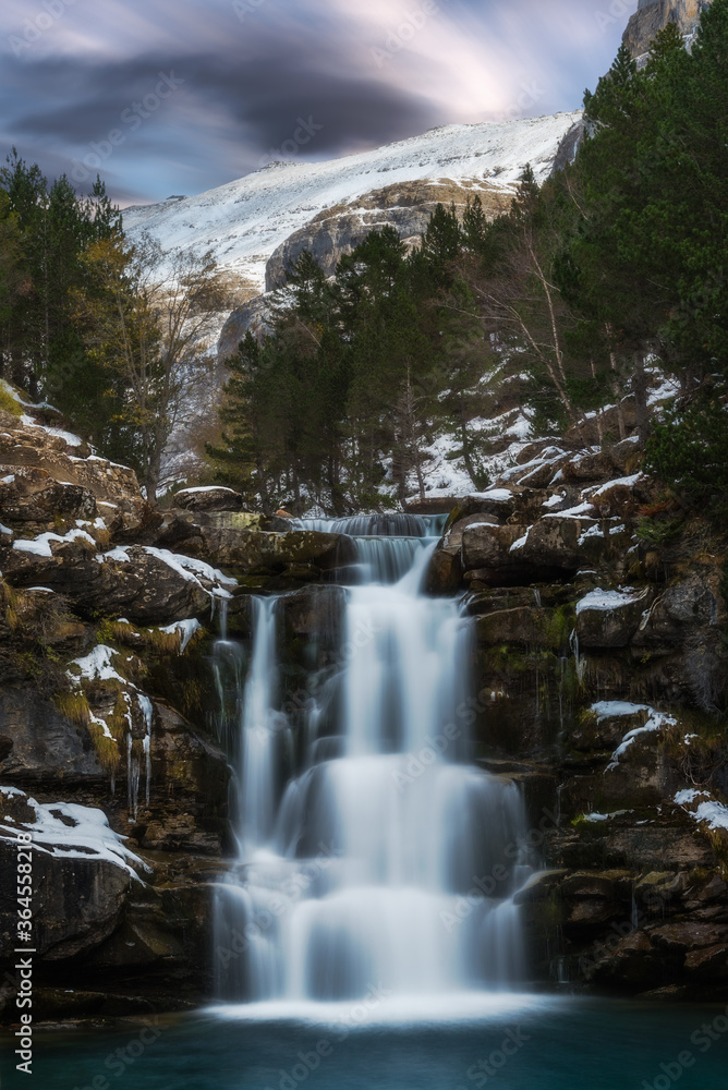 Scenic incredible view of small mountain cold waterfall flowing through snowy cliffs surrounded by coniferous forest on winter Scenic incredible view of small mountain cold waterfall flowing through snowy cliffs surrounded by coniferous forest on winter