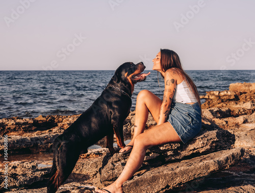 Side view of trendy tattooed woman sitting on stone and kissing Rottweiler dog while spending time on rocky seashore photo