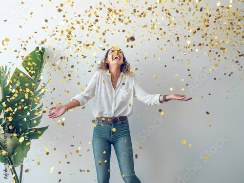 Beautiful young woman in stylish outfit laughing and looking up while standing under¬†falling golden confetti against white wall photo