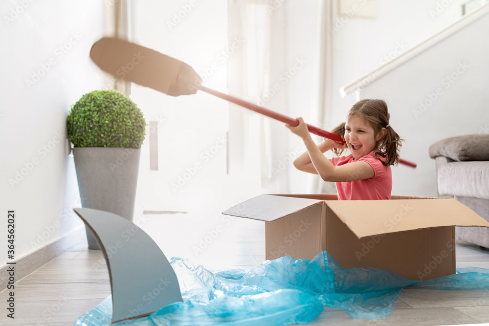 Playful preteen kid sitting in carton box at home and pretending to be sailor with paddle in boat screaming and pointing with finger on shark swimming beside